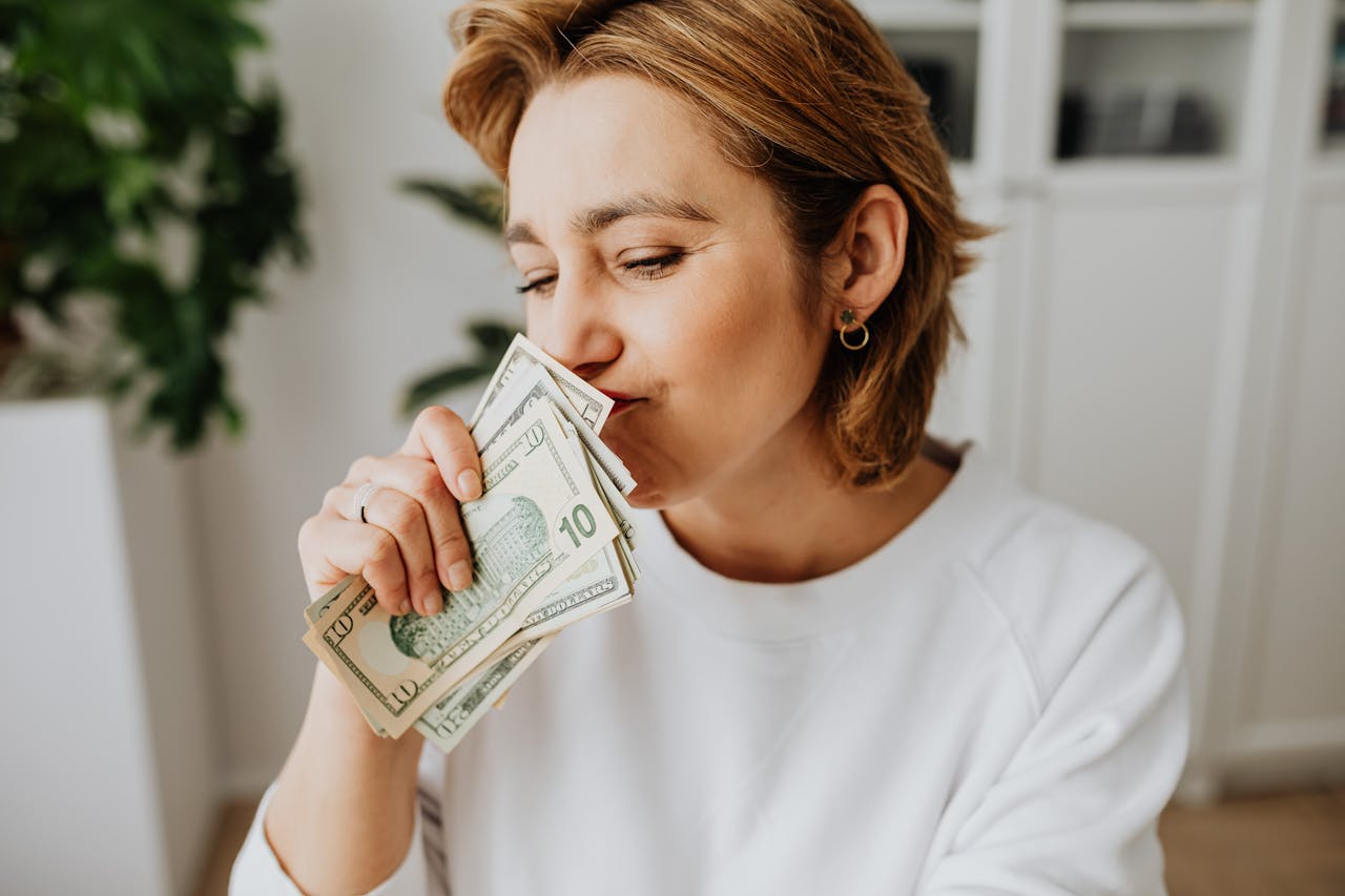 A woman joyfully smelling a handful of US dollar bills indoors.