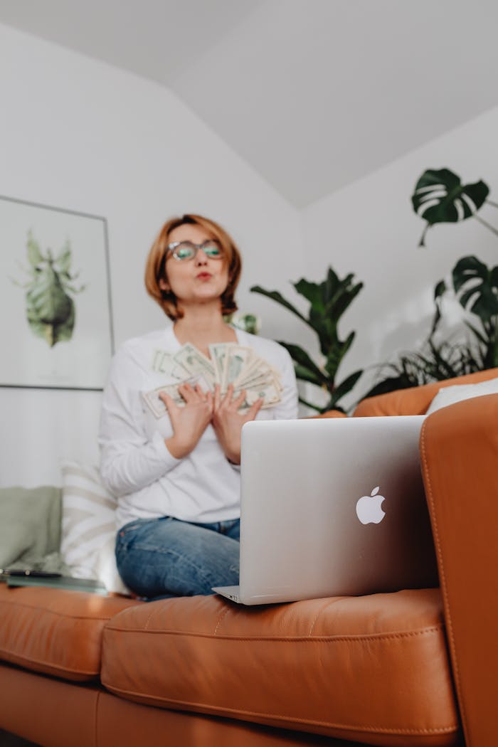 Woman holding cash and sitting on a sofa with a laptop indoors.